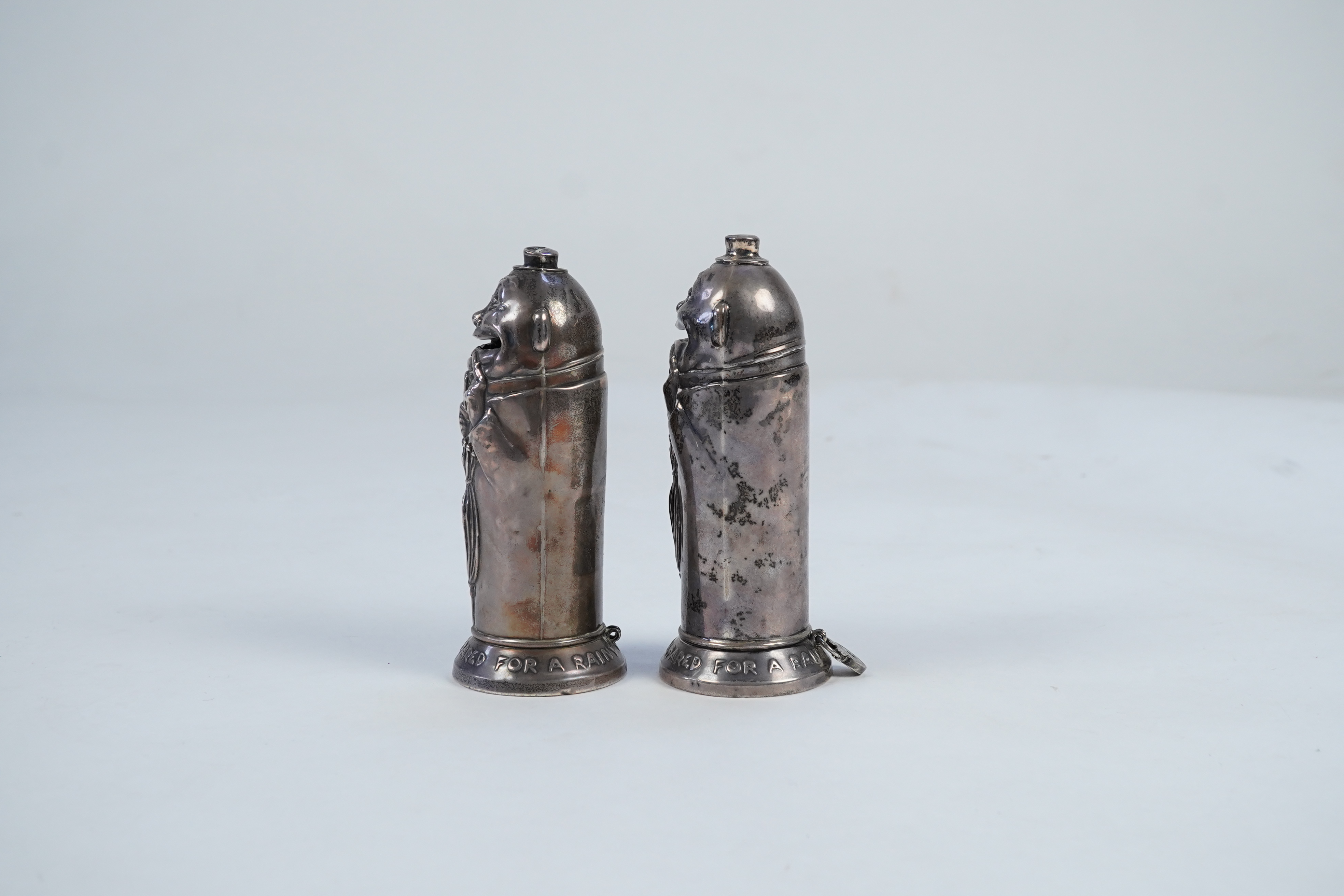 A matched pair of George V novelty silver money boxes, each cylindrical box embossed as a banker with an umbrella, the circular base inscribed 'Always be prepared for a rainy day'